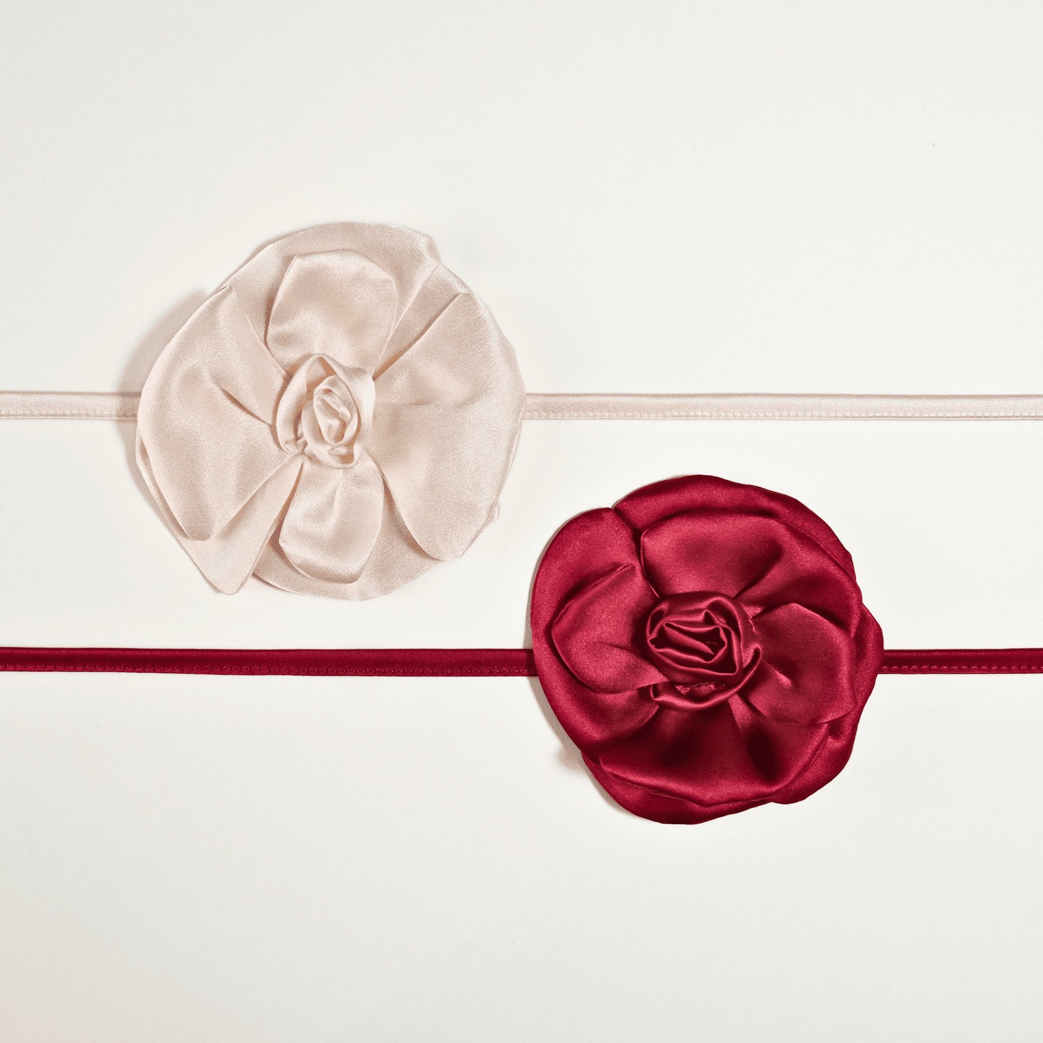 Due collane choker in seta della Alkira Collection con fiore artigianale cucito a mano, nei colori bianco cipria e burgundy, fotografate distese su sfondo bianco.